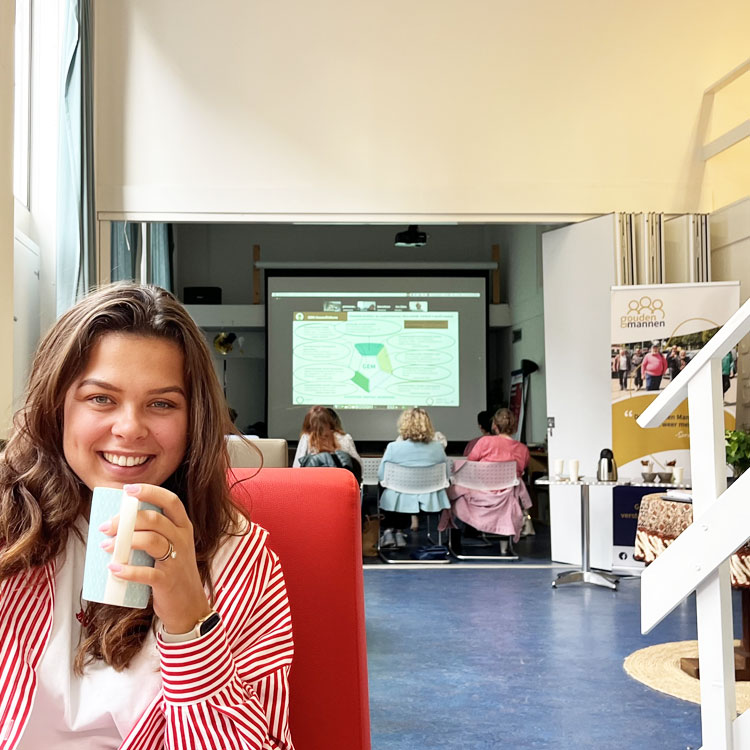 Aardig glimlachende vrouwelijke adviseur, die je aankijkt, zittend op een bank, met een pakje drinken. Ze heeft een rood gestreept bloesje aan.